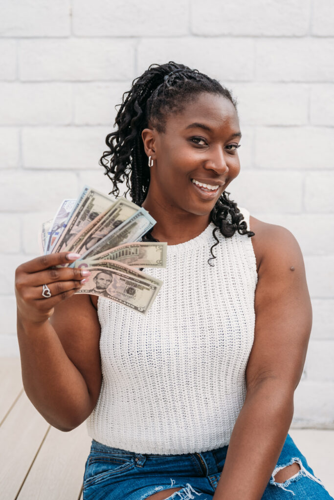 Smiling woman in a white sleeveless top and ripped jeans holds a fan of various currencies, conveying happiness and financial success. Light brick wall backdrop.