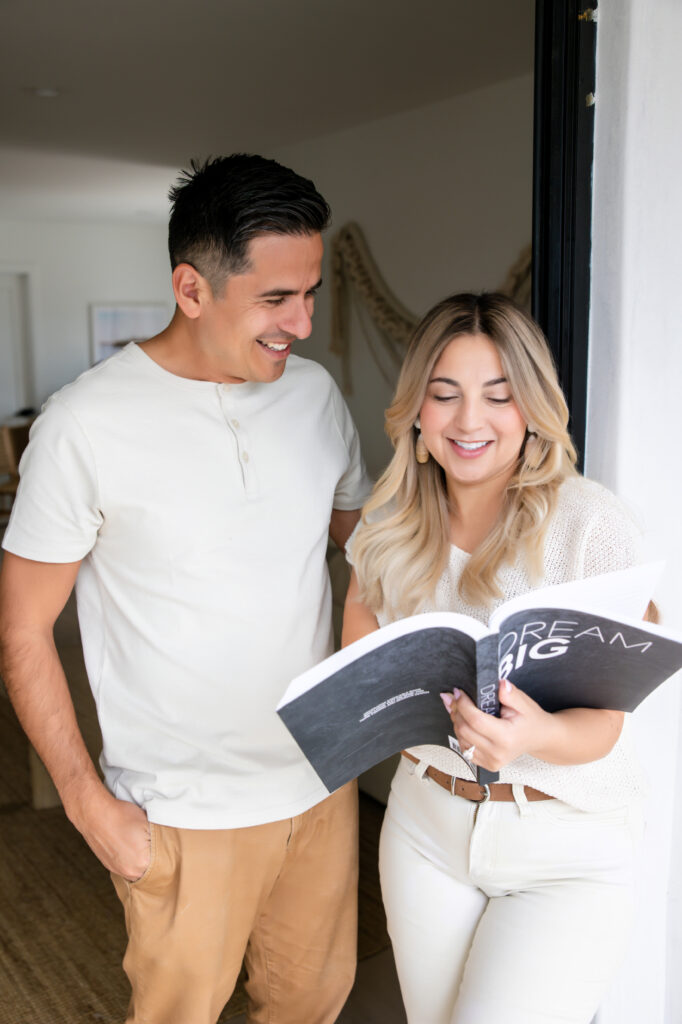 A smiling woman and man stand together indoors, looking at a book titled "Dream Big." They appear happy and relaxed in a bright, cozy space.