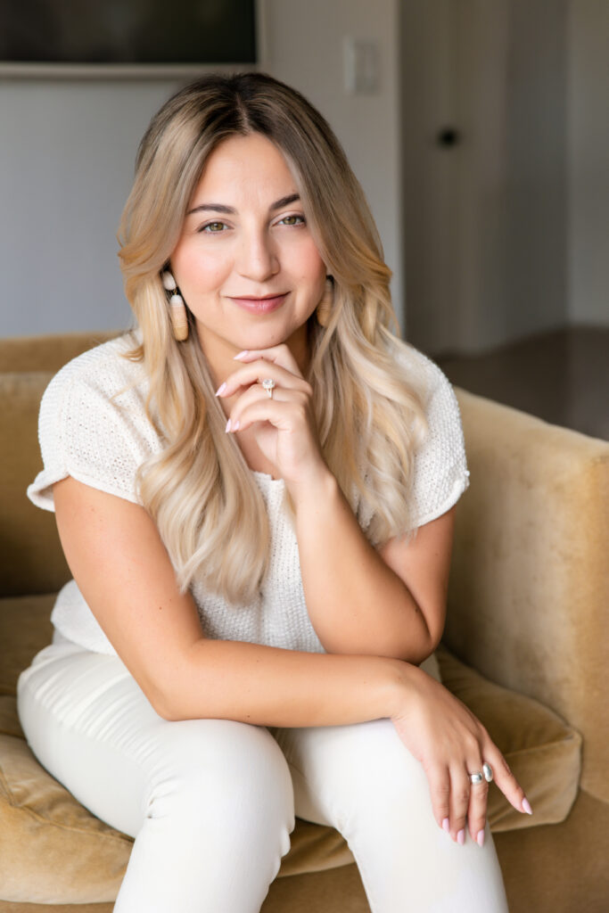 Woman with long blonde hair sits on a yellow couch, wearing a white knit top and pants. She smiles softly, exuding confidence and warmth.