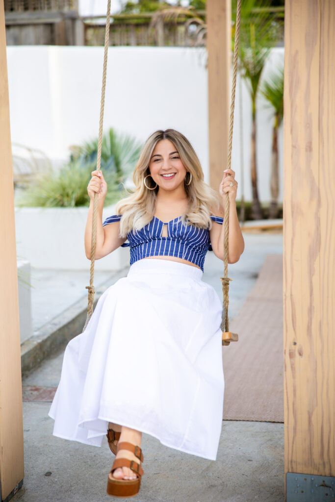 Woman in a blue striped top and white skirt smiles while sitting on a wooden swing. She wears hoop earrings, conveying a joyful and relaxed mood.