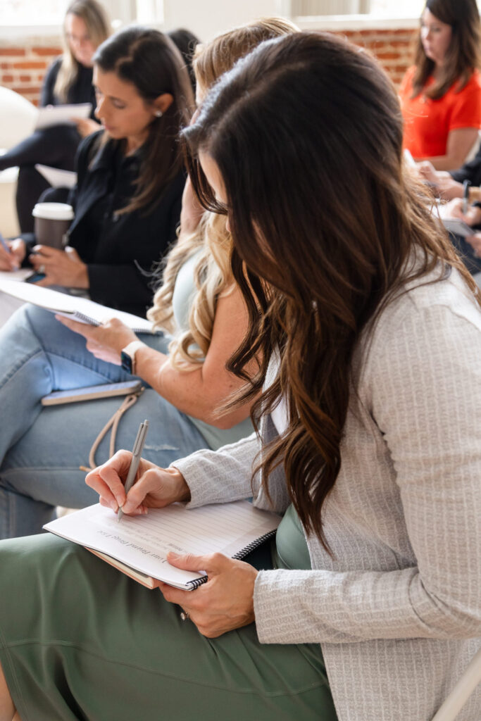 Women attentively writing notes in a notepad during a meeting or workshop. The atmosphere appears focused and collaborative.