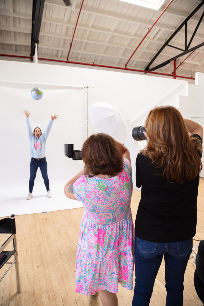 A photoshoot scene with a person joyfully jumping, tossing a globe in a bright studio. Two photographers capture the moment, conveying excitement and creativity.