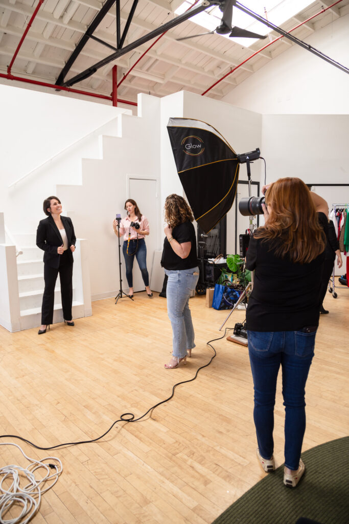 A photoshoot setup with a woman in a black suit smiling, posing near white stairs. Three photographers adjust equipment and take pictures in a bright studio.