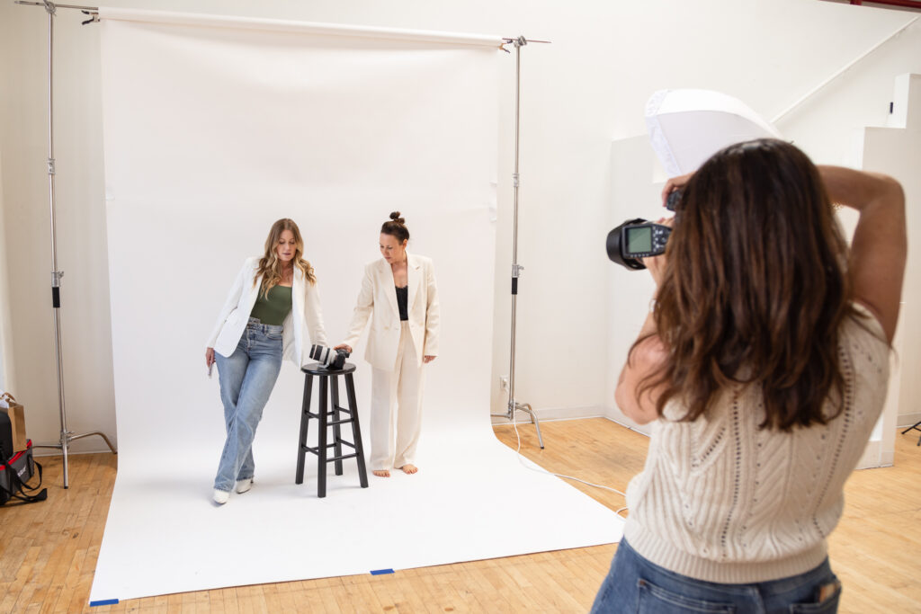 Photographer captures two women in a studio. One leans on a stool; both wear white blazers. Bright, airy atmosphere with a wooden floor.