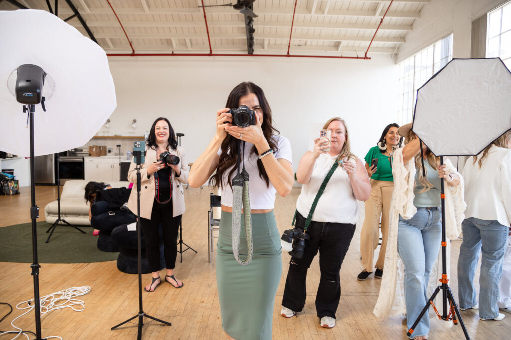 A group of women in a bright studio, smiling and taking photos. Some hold cameras, others adjust lighting equipment. The mood is lively and creative.