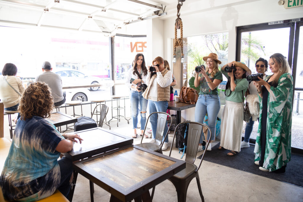 A group of women in casual outfits with cameras enthusiastically take photos of a woman sitting at a café table. Bright, lively atmosphere, with natural light.