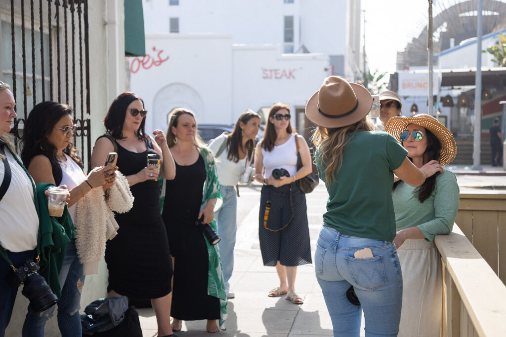 A group of women, some holding cameras and iced coffees, gather outdoors with casual attire. One adjusts another's hat, expressing a relaxed and friendly vibe.
