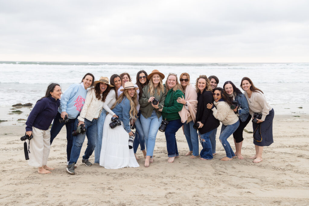 A joyful group of women photographers poses barefoot on a beach, each holding a camera. They are smiling and dressed casually with the ocean in the background.