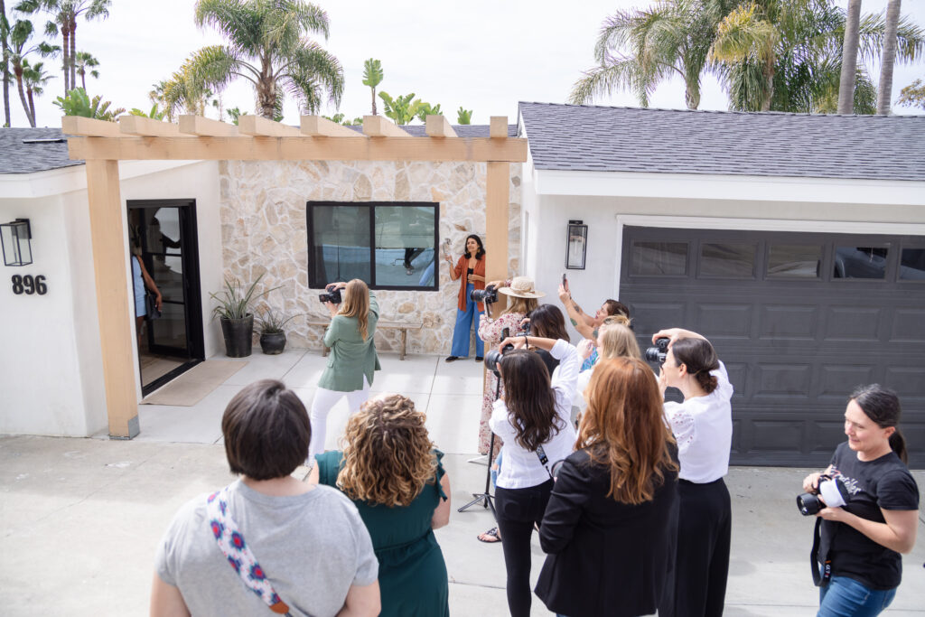 A group of people with cameras gather outside a modern house with a stone facade and black garage door, capturing a woman posing under a pergola. Palm trees are in the background, creating a lively and engaged atmosphere.