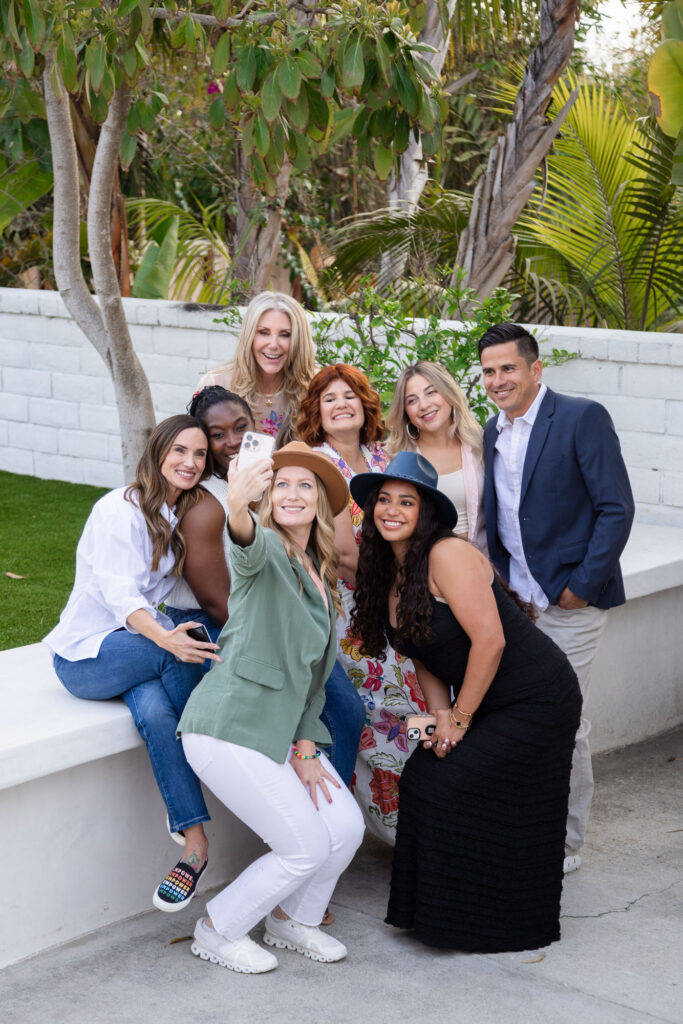 A group of eight people gather outside, smiling and taking a selfie. They're casually dressed, surrounded by greenery, creating a joyful, relaxed atmosphere.