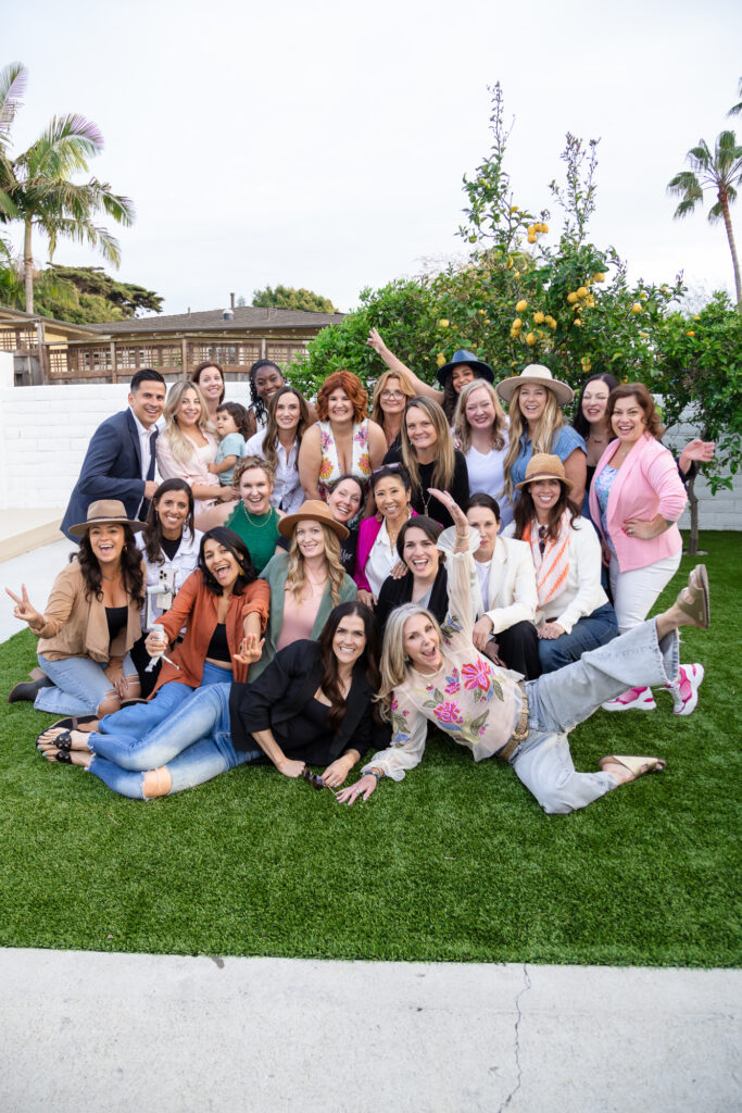 A diverse group of smiling people gathered outdoors on a lawn, posing cheerfully for a photo. Some wear hats, with lush greenery and palm trees in the background.
