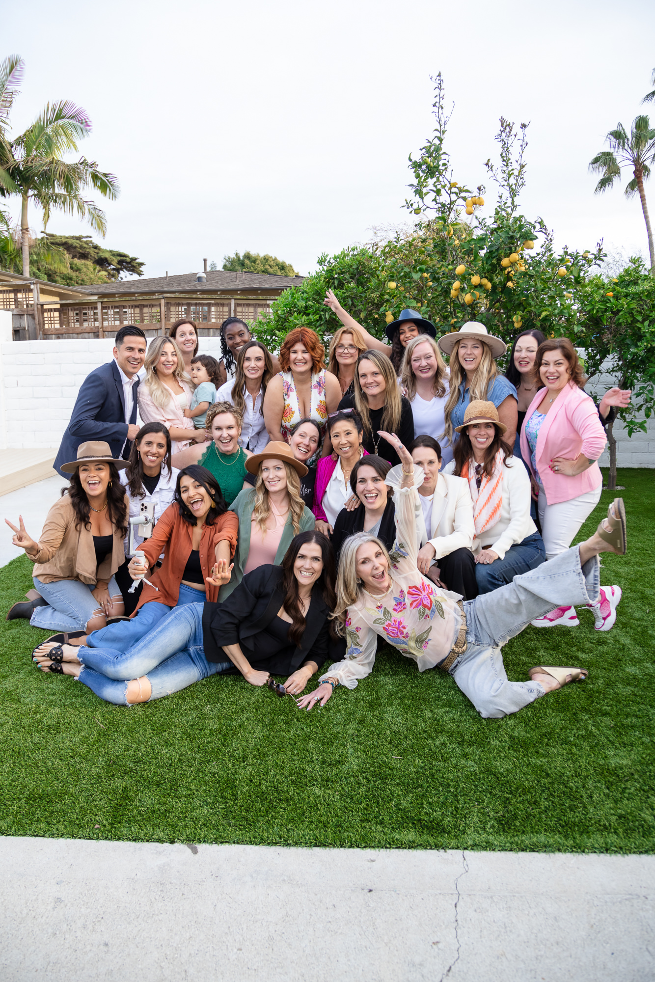 A joyful group of people poses outdoors on grass, some wearing hats. A cheerful atmosphere with palm trees and a lemon tree in the background.