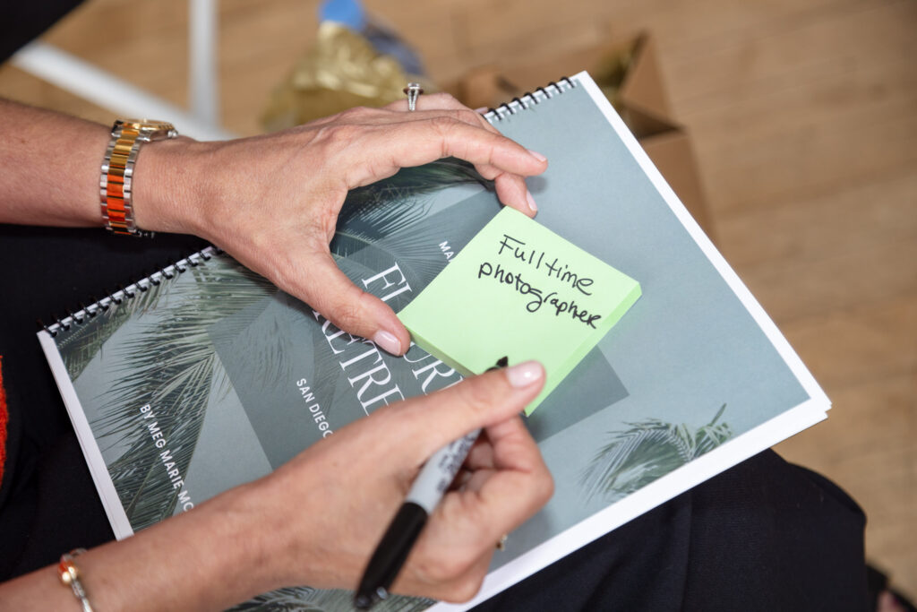 Hands with a silver watch hold a notebook and a green sticky note reading "Full-time photographer." A black marker rests in the right hand.