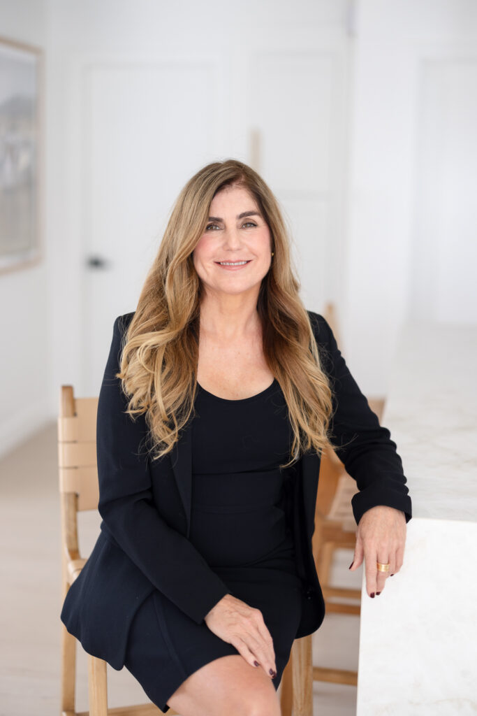 Woman with long, wavy hair in a black dress sits on a white couch, smiling warmly. Large green plant and minimal decor create a modern, serene vibe.