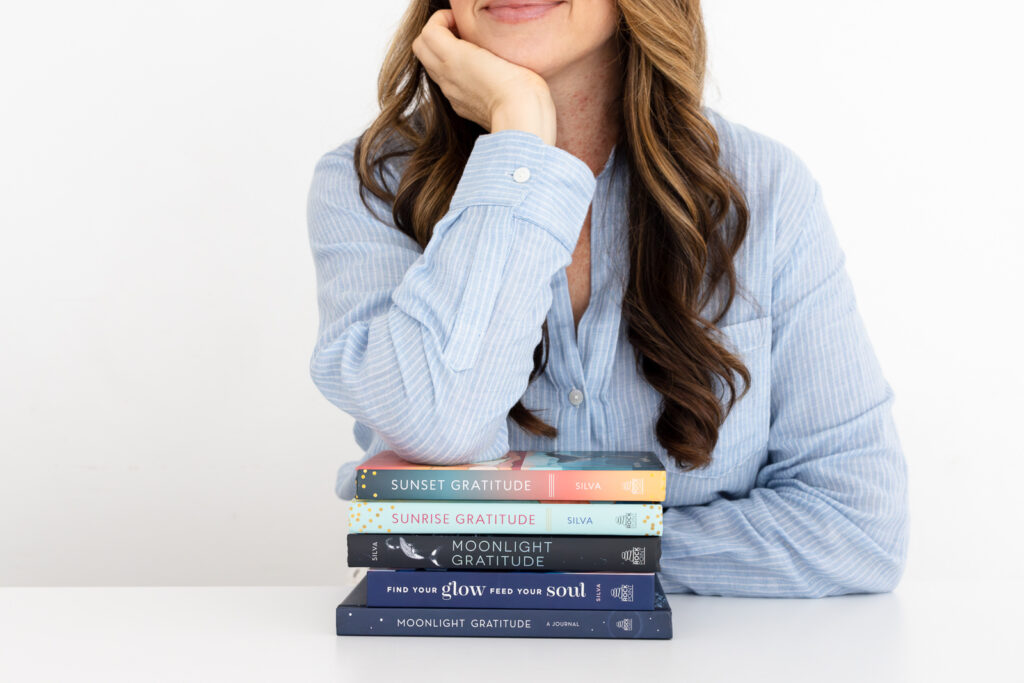 A woman in a blue shirt rests her chin on her hand beside a stack of books titled "Sunset Gratitude," "Sunrise Gratitude," "Moonlight Gratitude," and others. The scene conveys tranquility and contemplation.
