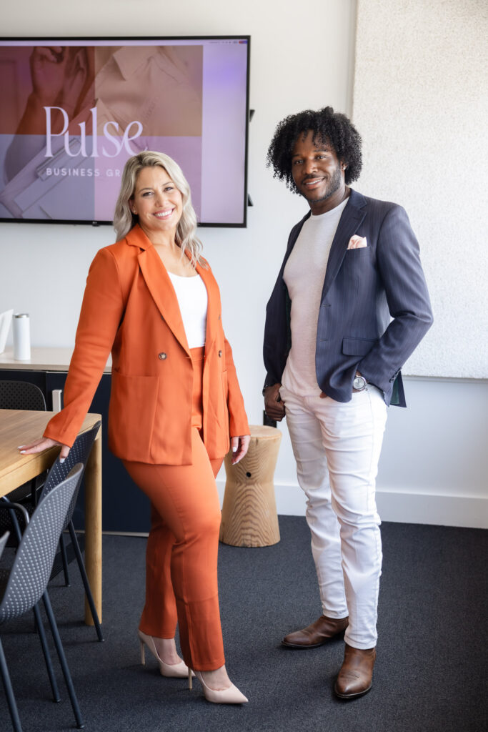 A woman in an orange suit and a man in a dark blazer and white pants stand smiling in a modern office setting, conveying a professional, friendly tone.