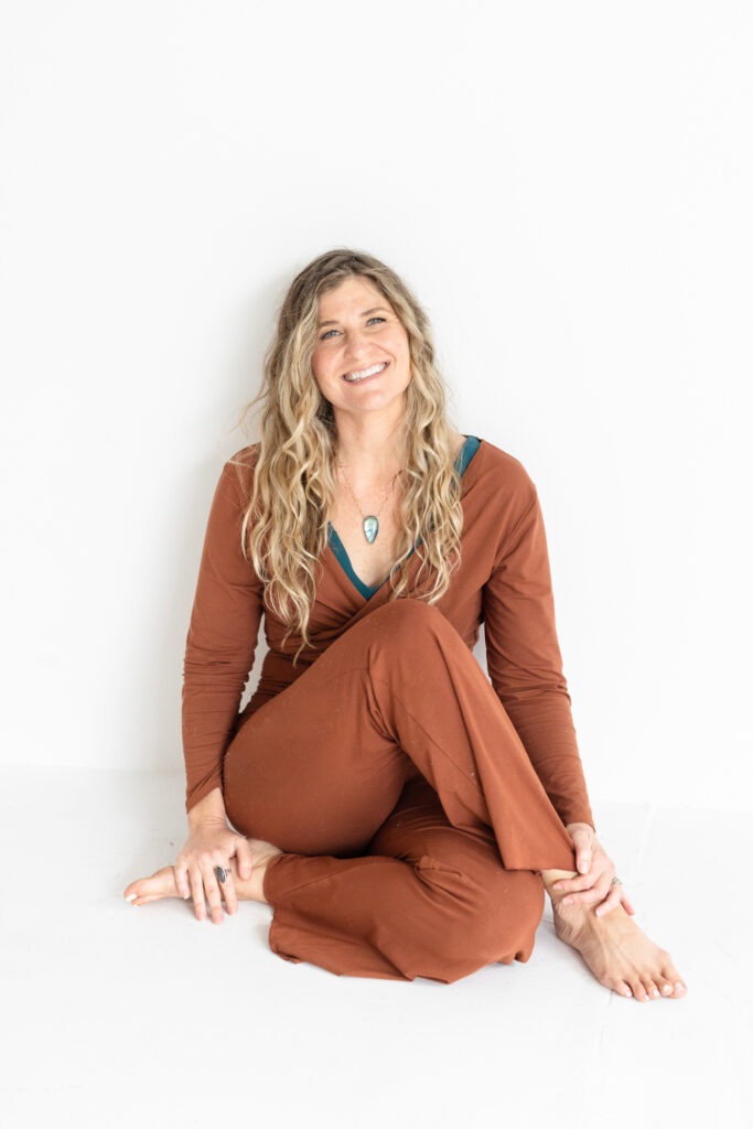 A woman with long, wavy hair sits cross-legged, smiling warmly. She wears a rust-colored outfit with a teal pendant. The background is a plain white wall, offering a serene and relaxed atmosphere.