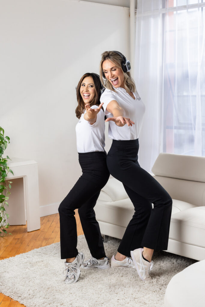 Two women in white tops and black pants joyfully pose together, one wearing headphones, in a bright room with a sofa and plants, conveying energy and happiness.