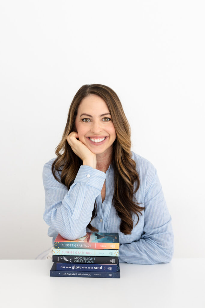 A woman in a light blue shirt smiles warmly, resting her chin on her hand. In front of her is a stack of colorful books against a plain white background.