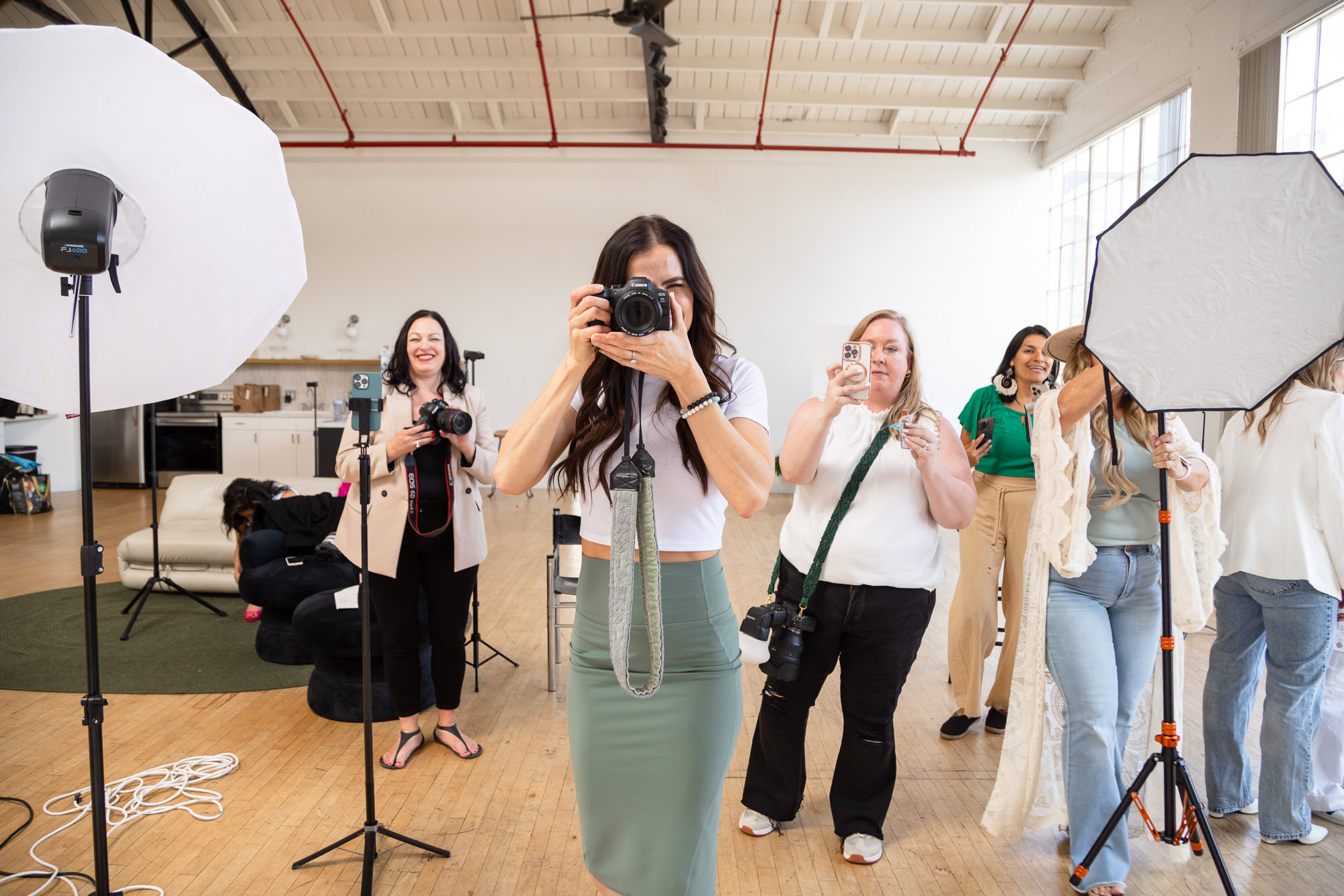 A group of seven women in a bright studio, holding cameras and posing mid-action. The ambiance is lively and creative, with photography equipment visible.