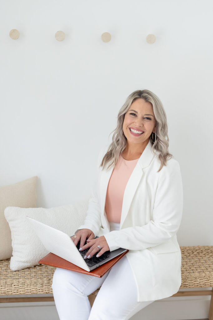 Smiling woman with wavy hair sits on a bench, wearing a white blazer and peach top, working on a laptop. Background is minimalist and bright.