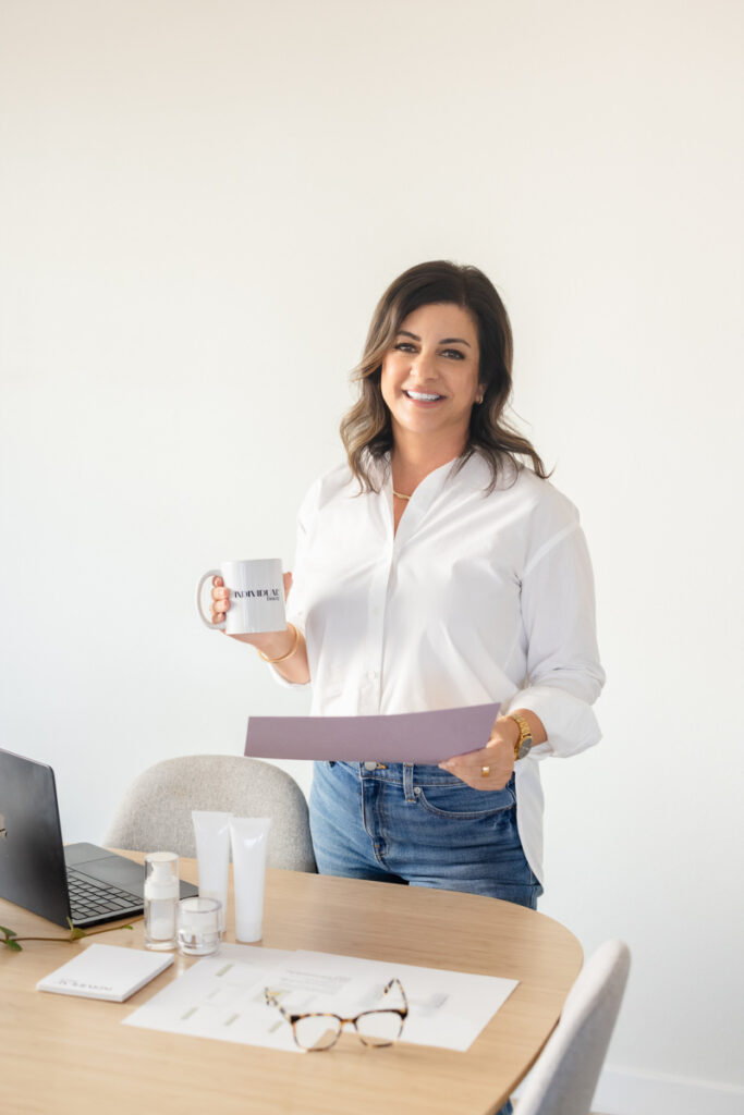 A woman in a white shirt and jeans smiles while holding a mug and document. She stands by a table with a laptop, papers, cosmetics, and glasses. Cozy, professional atmosphere.