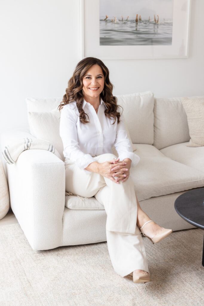 A woman with wavy hair, seated on a white sofa, smiles warmly. She's wearing a white blouse and pants, creating a calm, welcoming ambiance. A serene ocean painting is in the background.