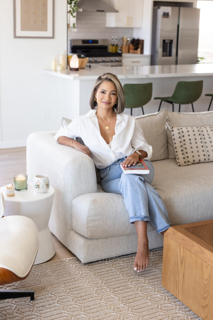 A woman sits on a light beige sofa in a modern, bright living room. She's wearing a white blouse and jeans, holding a book. The background features a sleek kitchen with green bar stools. Candles on the table add a cozy touch.