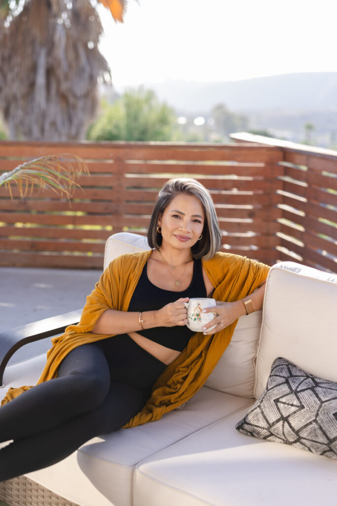 A woman in a mustard cardigan relaxes on an outdoor couch, holding a mug. She smiles confidently, with a wooden fence and trees in the background.