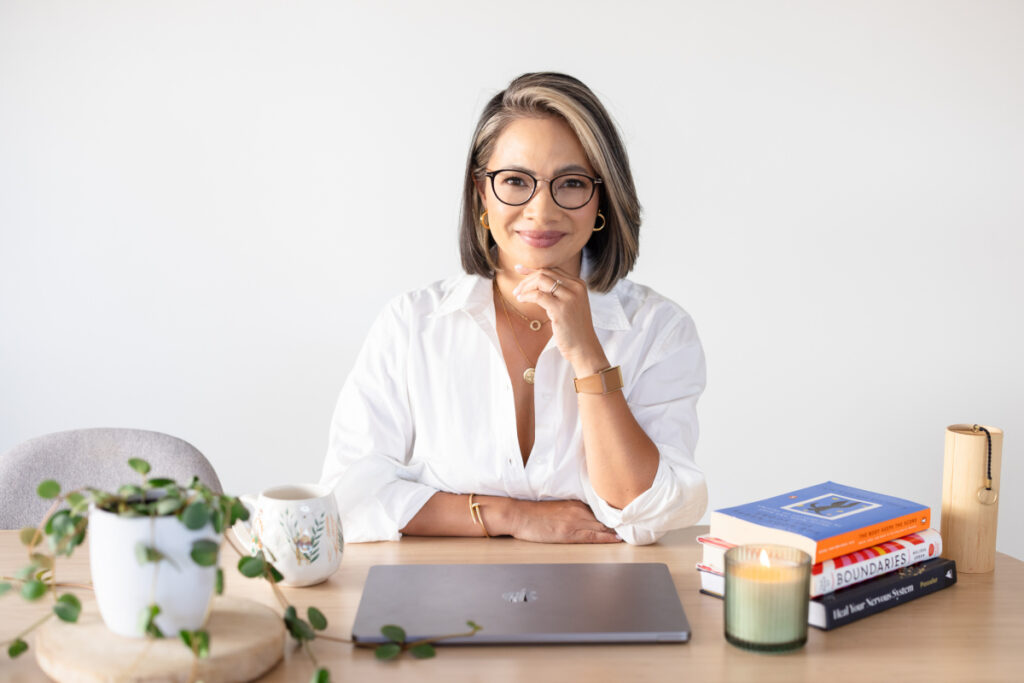 A confident woman with glasses in a white shirt sits at a desk with a laptop, potted plant, candle, and stack of books, smiling at the camera.