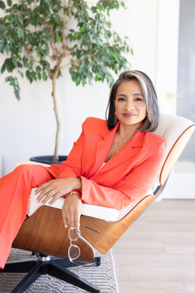 A woman in a bright orange suit relaxes in a stylish chair, holding glasses. She smiles confidently. A lush green plant is visible in the background.