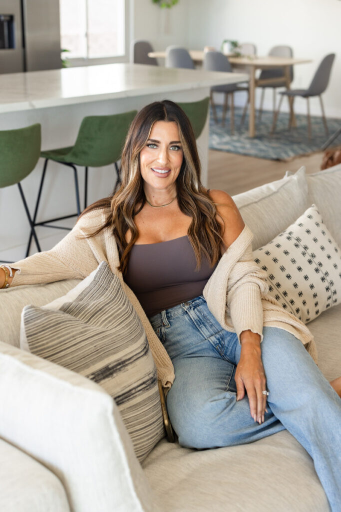 Woman with long brown hair in a beige cardigan and jeans sitting on a sofa, smiling warmly. Behind her, a modern kitchen and dining area are visible.