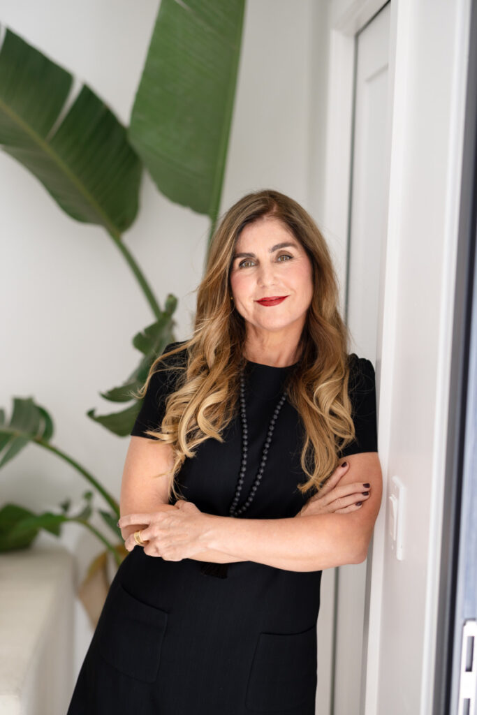 Woman with long wavy hair and black dress leans against a white wall, arms crossed, smiling confidently. Green plants in the background add a fresh touch.