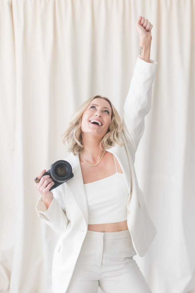 A joyful person in a white suit, holding a camera, raises an arm triumphantly against a cream curtain backdrop, conveying celebration and success.