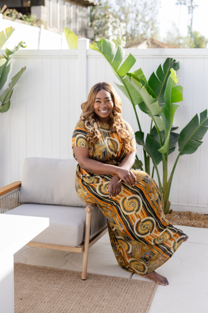 A woman with long brown hair and a black sleeveless top smiles joyfully outdoors, conveying a cheerful and relaxed vibe. Light-colored house in the background.