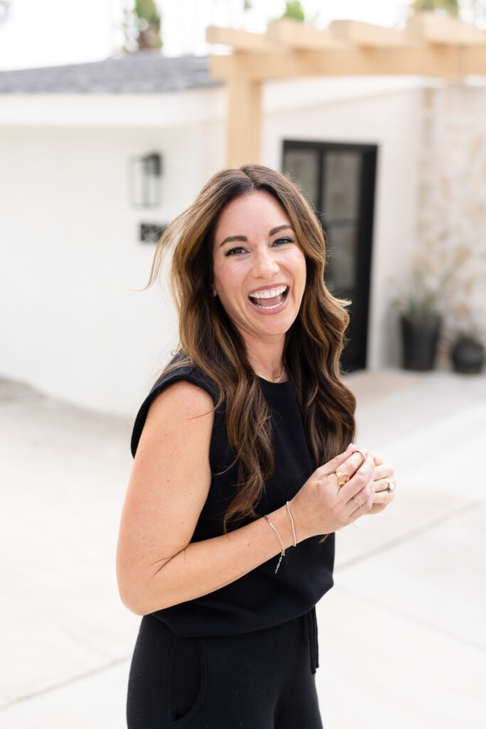 A woman with long brown hair and a black sleeveless top smiles joyfully outdoors, conveying a cheerful and relaxed vibe. Light-colored house in the background.