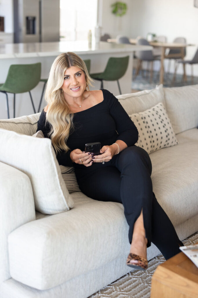 A woman with long blonde hair in a black outfit sits on a beige sofa, holding a phone. Modern living room with green chairs in the background. Cozy and relaxed atmosphere.