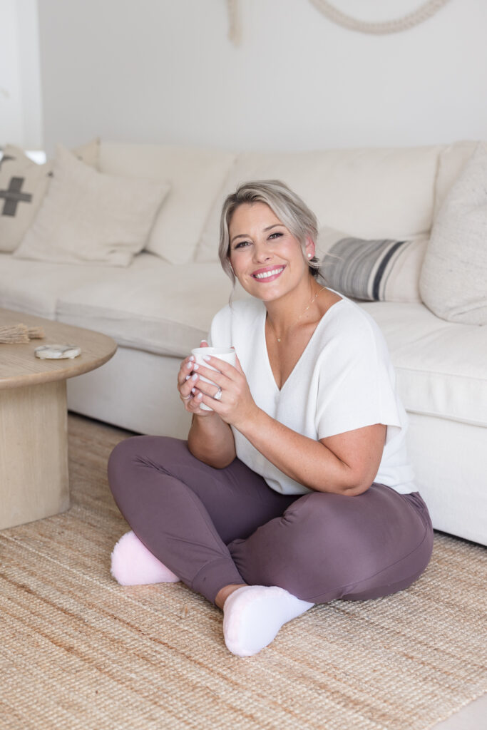 A woman with short blonde hair sits cross-legged on a beige rug in a cozy living room, holding a mug and smiling warmly. The background features a cream sofa with pillows.
Meg Marie Photography | San Diego