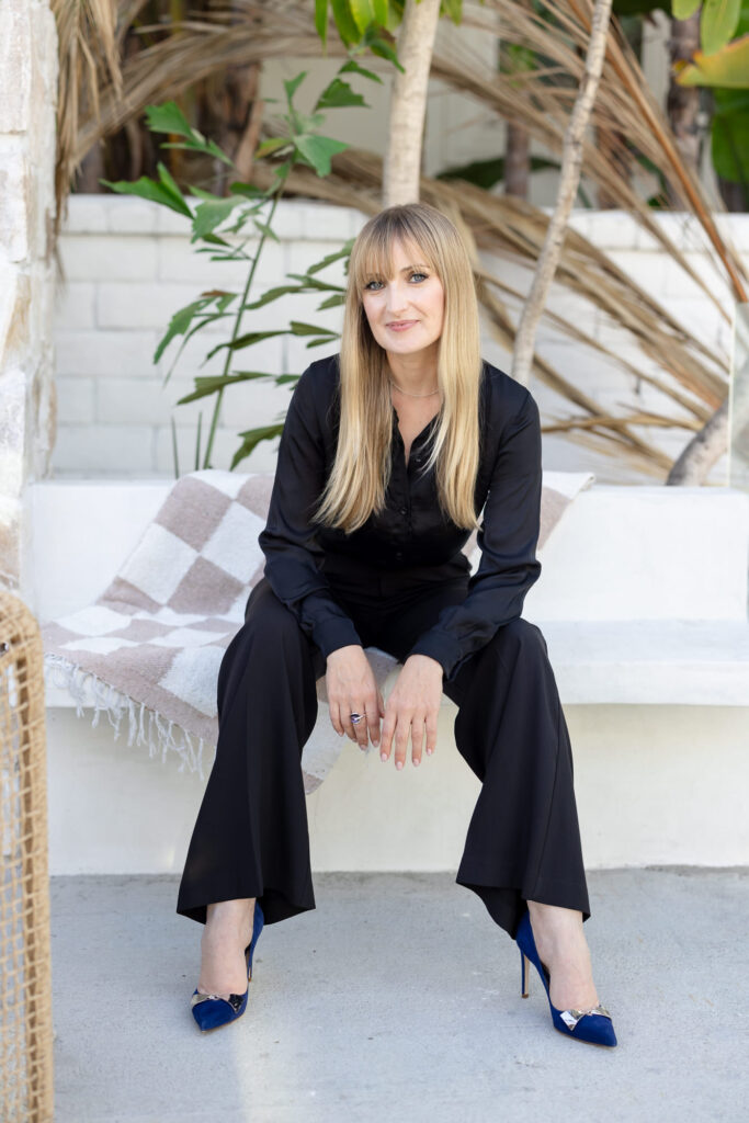 A woman with long blonde hair and a black outfit sits confidently on a white bench, surrounded by green plants. She wears blue high heels; the scene is calm and stylish.
Meg Marie Photography | San Diego