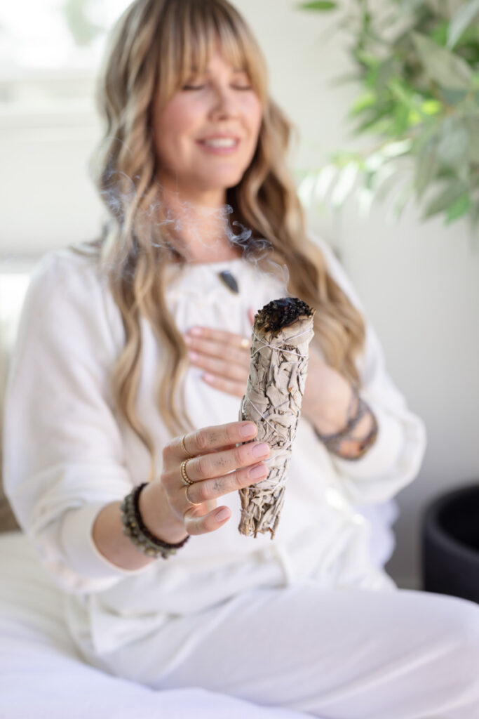 A woman with long hair holds a smoldering sage stick, wearing white and sitting on a bed. She appears relaxed and content, with plants nearby.
Meg Marie Photography | San Diego