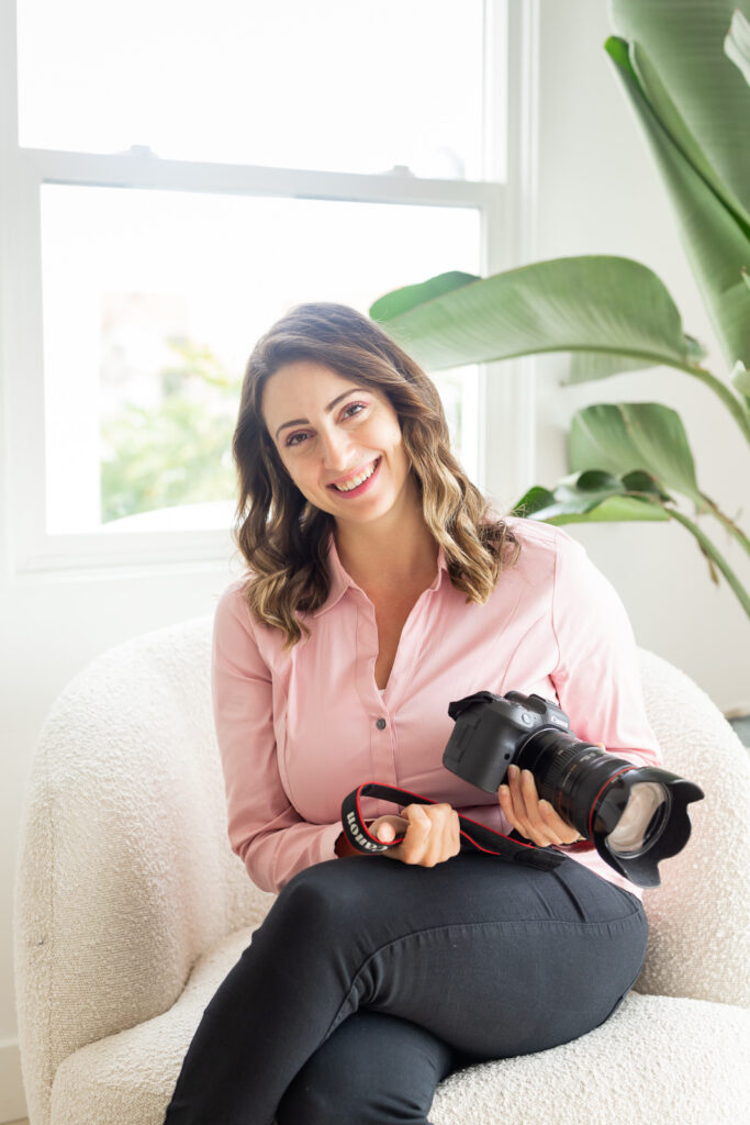 Smiling woman in a pink blouse holds a camera, sitting on a white chair near a window. Lush green plant in the background, bright and warm atmosphere. | Meg Marie Photography