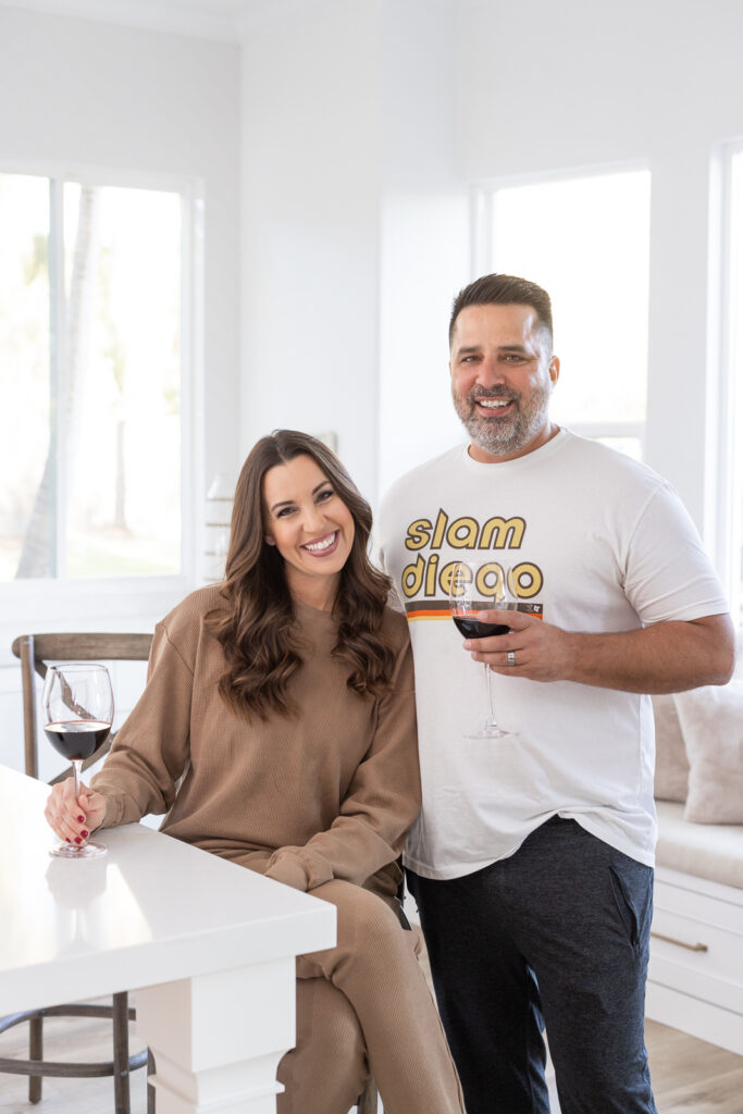 A smiling woman in a brown outfit and a man in a "Slam Diego" T-shirt stand in a bright kitchen, each holding a glass of red wine. The atmosphere is warm and relaxed. | Meg Marie Photography