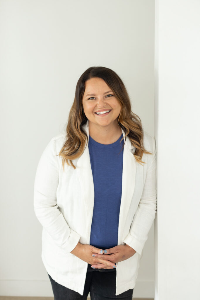 Smiling woman with long brown hair leans against a white wall. She wears a blue shirt and white jacket, exuding warmth and friendliness in a minimalist setting. | Meg Marie Photography