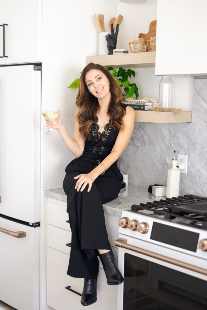 A woman in black lace top and pants sits on a kitchen counter, holding a glass. The modern kitchen has marble backsplash and wooden shelves, creating a cozy ambiance. 