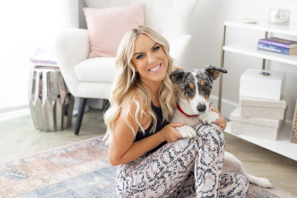 Smiling woman with long blonde hair sits on a patterned rug, hugging a dog in a bright room. Cozy atmosphere with soft chair, books, and decorative items. | Meg Marie Photography