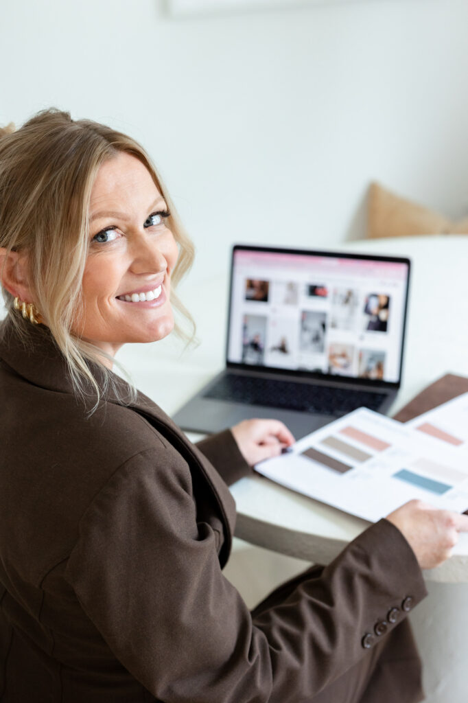 Smiling woman in a brown blazer sits at a table with a laptop displaying images. She's holding a color swatch book, exuding a professional, creative vibe. | Meg Marie Photography