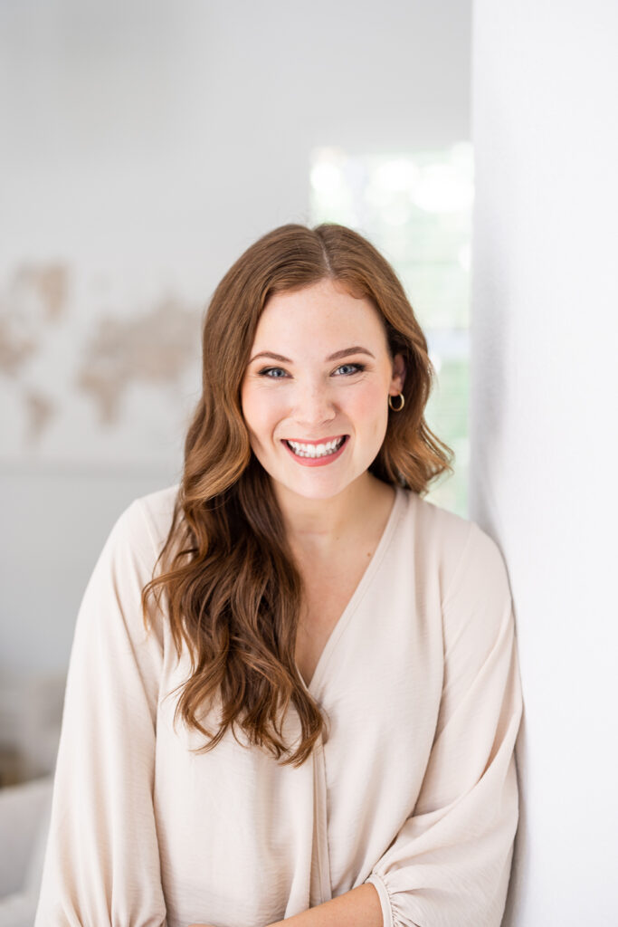 A woman with long, wavy hair and wearing a light beige top smiles warmly at the camera. The background is blurred, showing soft natural light, creating a cozy and friendly atmosphere. | Meg Marie Photography