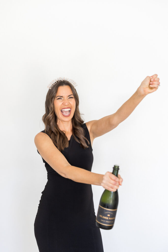 Smiling woman with long hair in a black dress opens a champagne bottle, raising her hand excitedly. She wears a tiara, conveying a celebratory mood. |   Meg Marie Photography