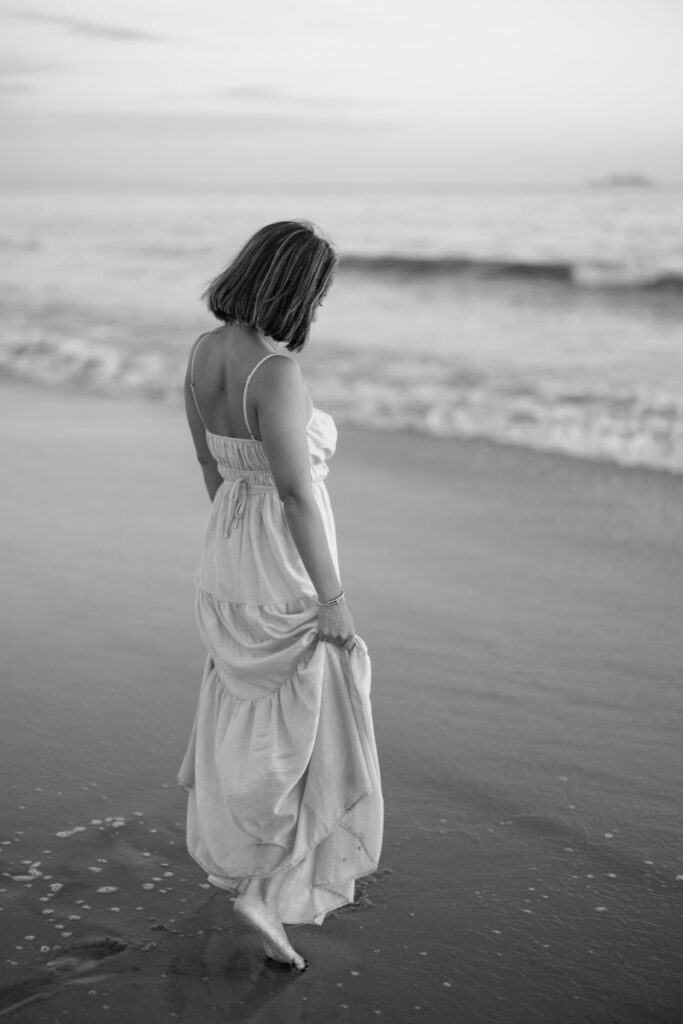 A woman in a flowing white dress walks barefoot on a serene beach, facing rolling waves. The black-and-white photo conveys a calm, reflective mood. | Meg Marie Photography