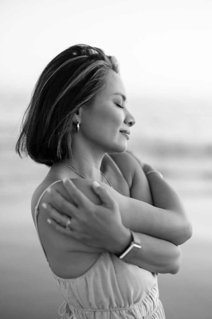 A woman with short hair, eyes closed, smiling softly, embraces herself at the beach. She wears a sleeveless top. The mood is peaceful and reflective. | Meg Marie Photography